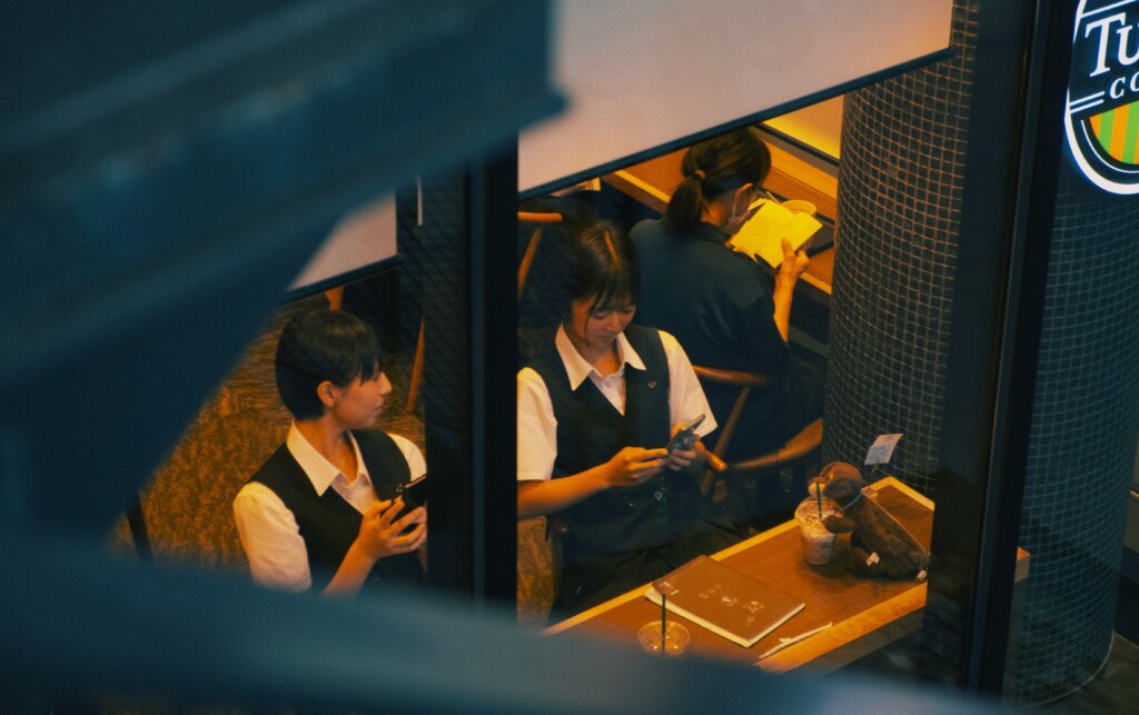 カフェでスマートフォンを操作する高校生たち。会話やSNSを通じて現代の言葉が広がる様子を象徴するシーン